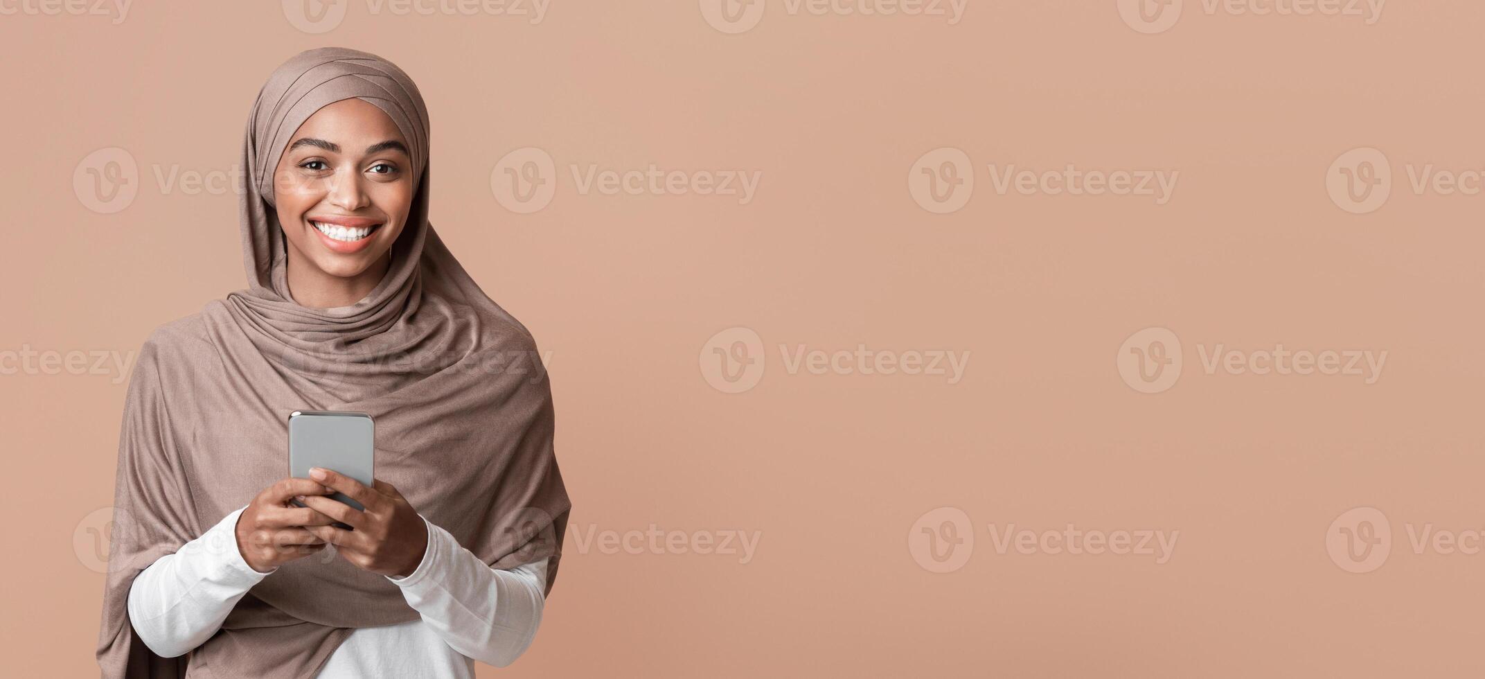 Modern Communication Concept. Portrait Of Cheerful Afro Muslim Girl In Hijab With Smartphone In Hands Looking At Camera, Yellow Background, Copy Space photo