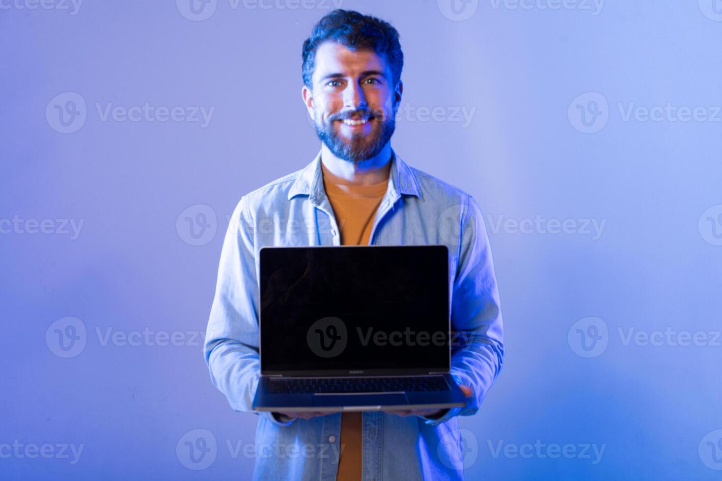 A smiling man wearing a light jacket stands indoors against a blue background, holding a laptop with black screen with both hands as he presents it toward the camera. photo