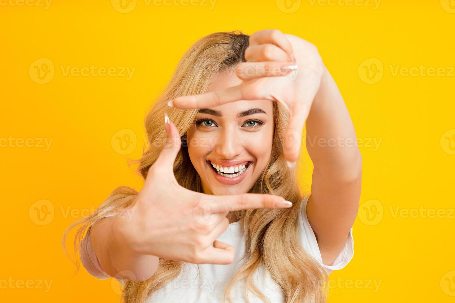 A young blonde woman with a bright smile is standing against a yellow background, making a frame with her hands. She is wearing a white t-shirt and has her hair flowing down her shoulders. photo