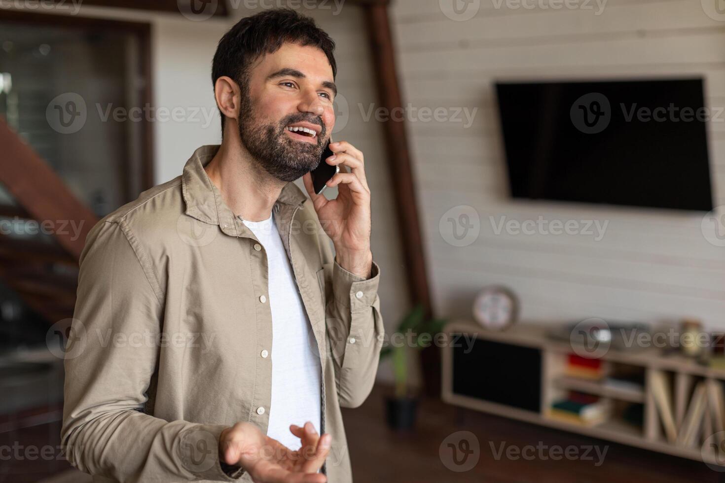 A man is engaged in a phone call while standing in a modern living room, holding a cell phone to his ear. The room is tastefully decorated with a sofa, coffee table, and plants, copy space photo