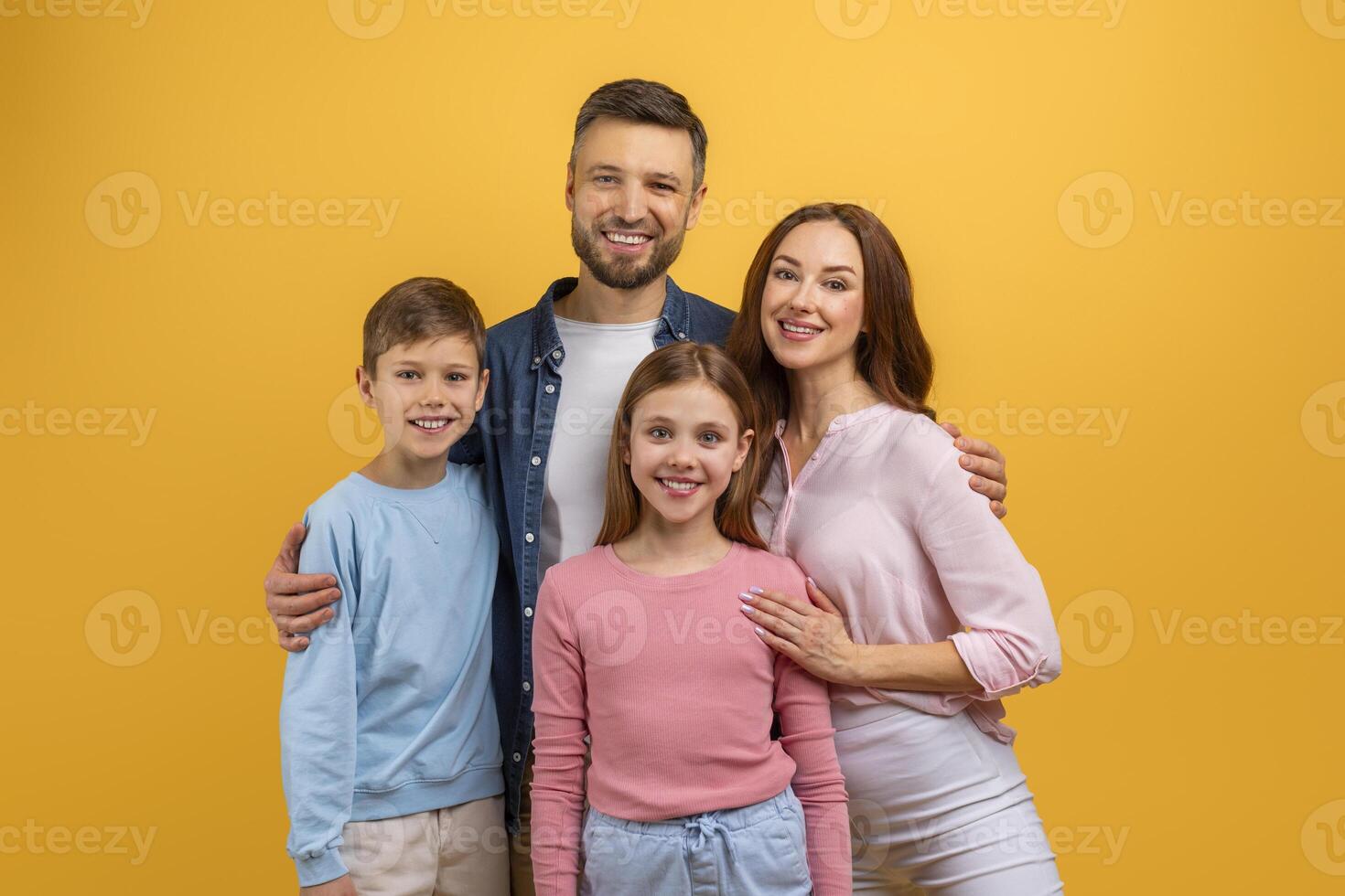 A family of four, consisting of two adults and two children, stands together in front of a vibrant yellow background, smiling and posing for a photograph, with the parents holding the children hands photo