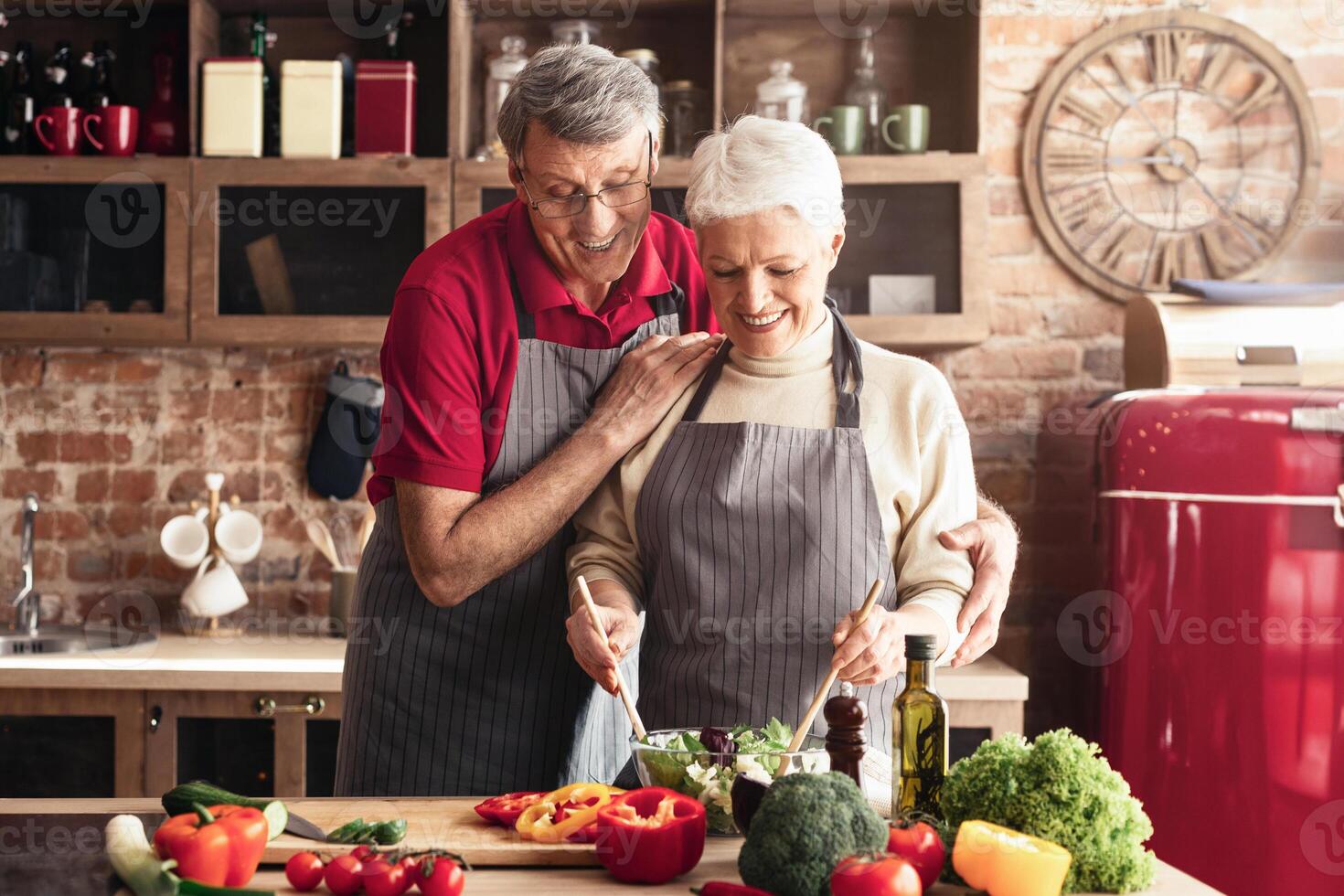 A senior woman with short, gray hair, wearing a white sweater and a gray striped apron, prepares a salad in a kitchen with a brick wall. She smiles as she stirs a bowl of greens with a wooden spoon photo