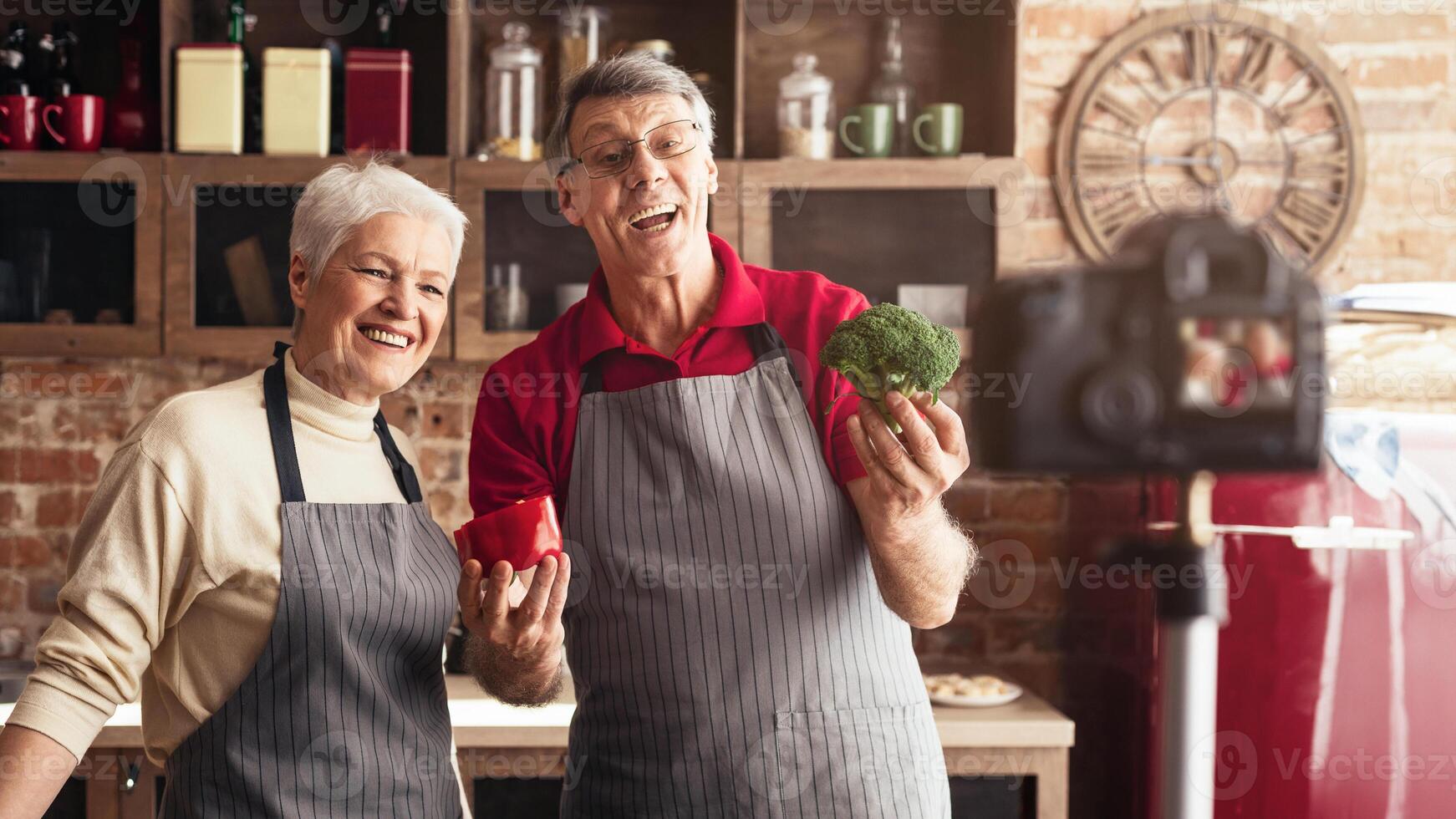 A DSLR camera is positioned in front of a senior couple in a kitchen. The camera lens is focused on the couple, who are both wearing aprons and appear to be cooking. photo