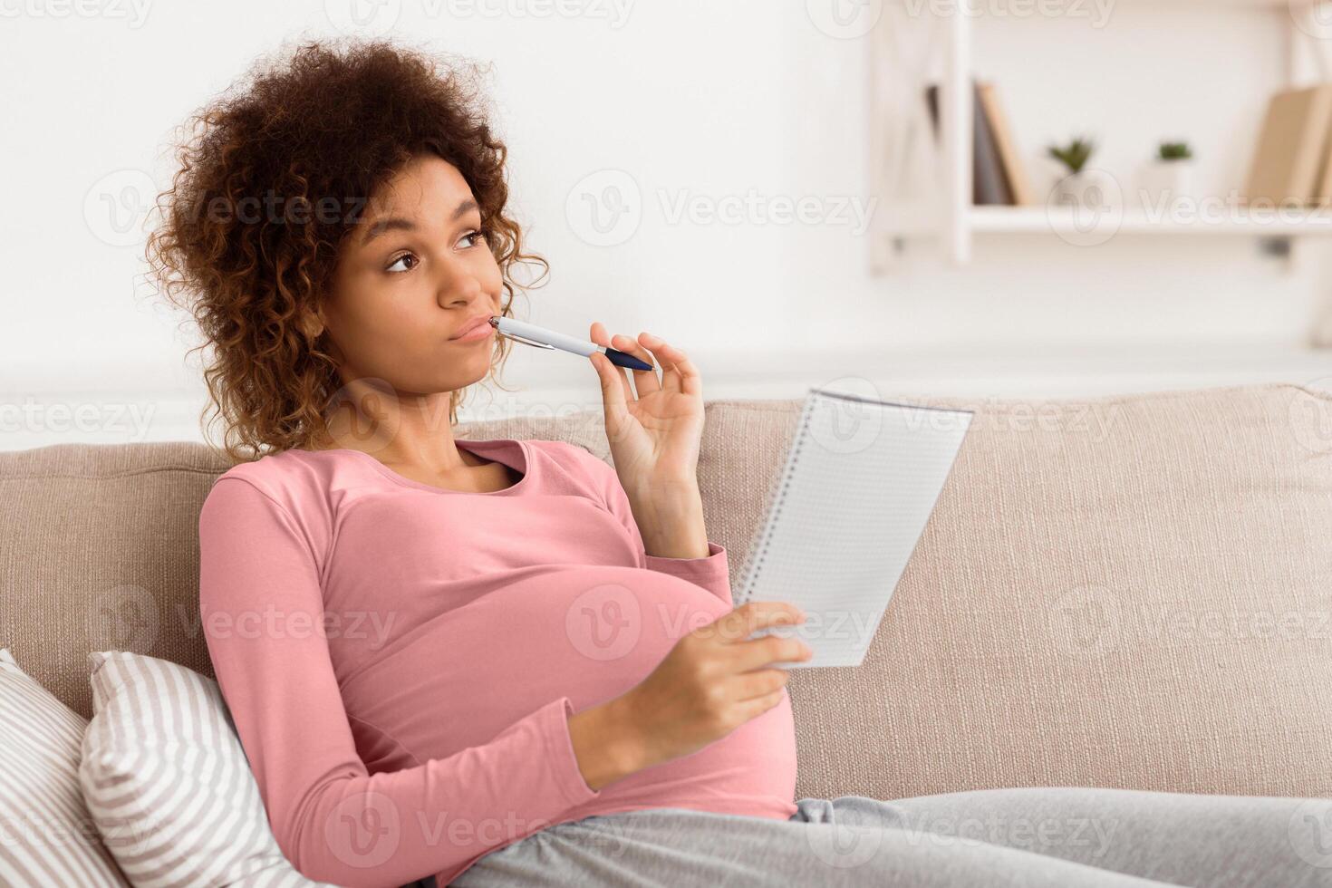Thoughtful pregnant lying on sofa with notebook and pen, making shopping list, free space photo