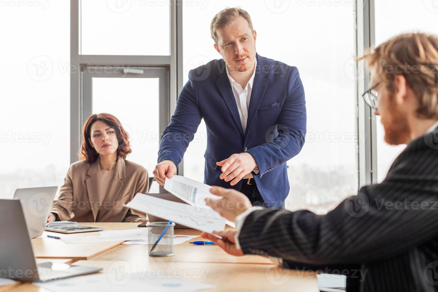 A businessman in a blue suit stands and hands documents to a seated colleague while another sits in the background, all gathered around a conference table in an office setting. photo