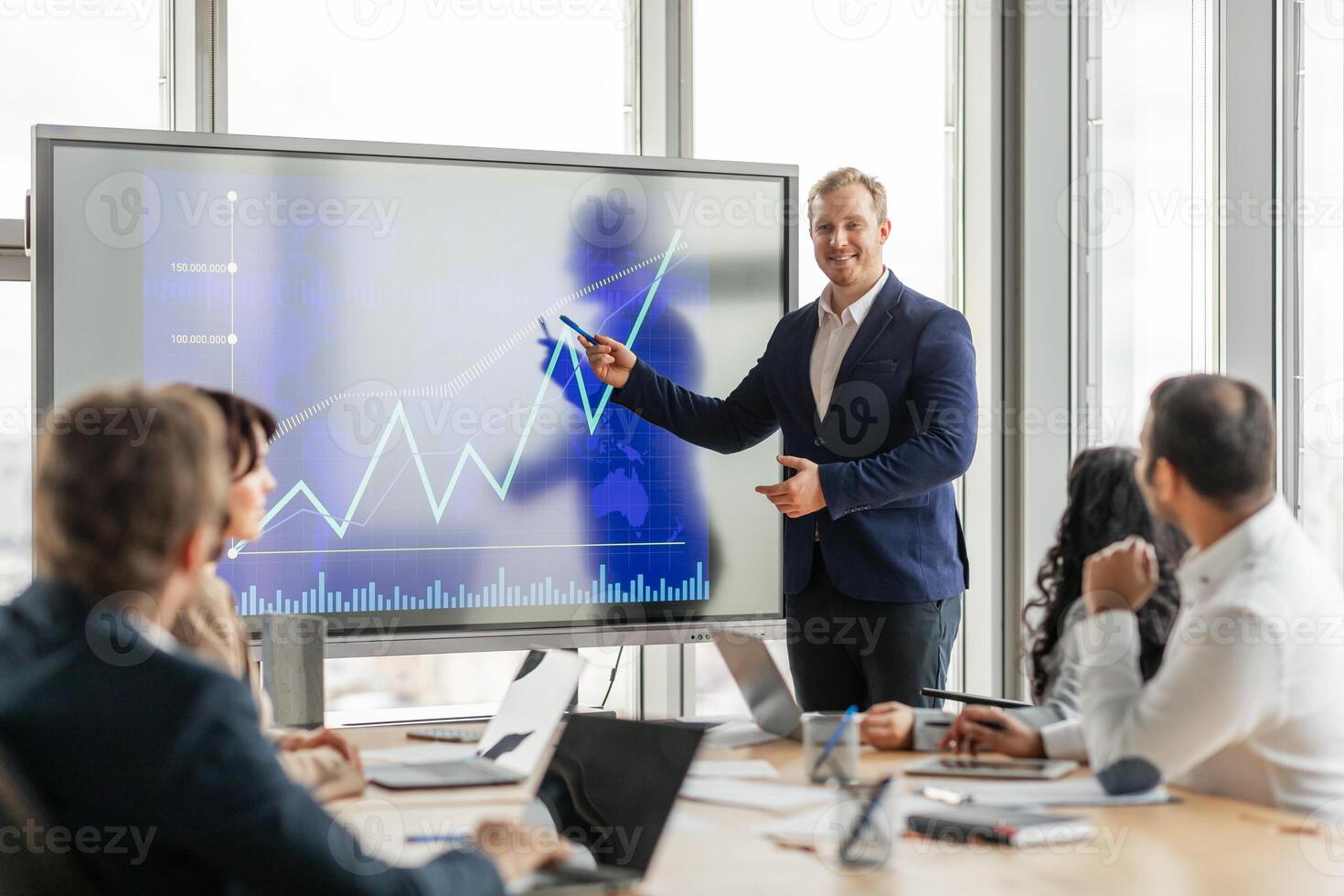 A businessman in a suit stands in front of a large screen, pointing to an upward trending graph. He is presenting to a group of colleagues who are seated at a conference table, listening attentively photo