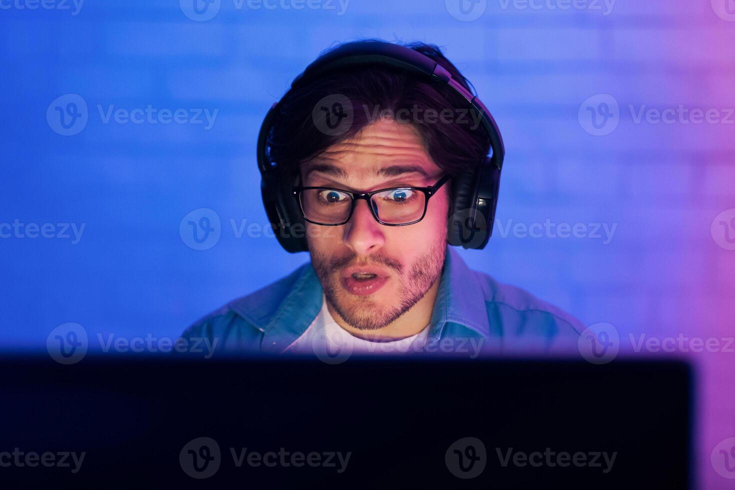 A young man with a shocked expression is seen staring intently at his computer screen while wearing headphones. The room is dimly lit with blue and purple hues, suggesting a late-night gaming session. photo