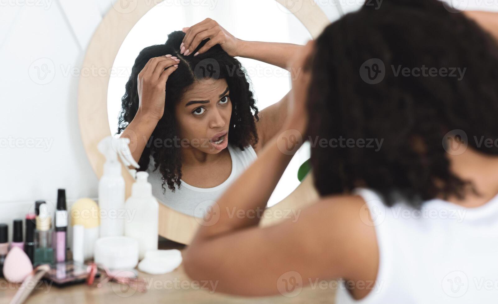 Dandruff Problem. Sad Black Woman Looking At Her Hair Roots With Flaky Scalp In Mirror, Sitting At Dressing Table, Over Shoulder View photo