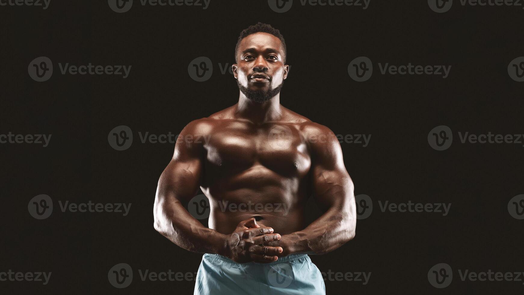 A muscular man poses with his hands clasped in front of him, against a black background. photo