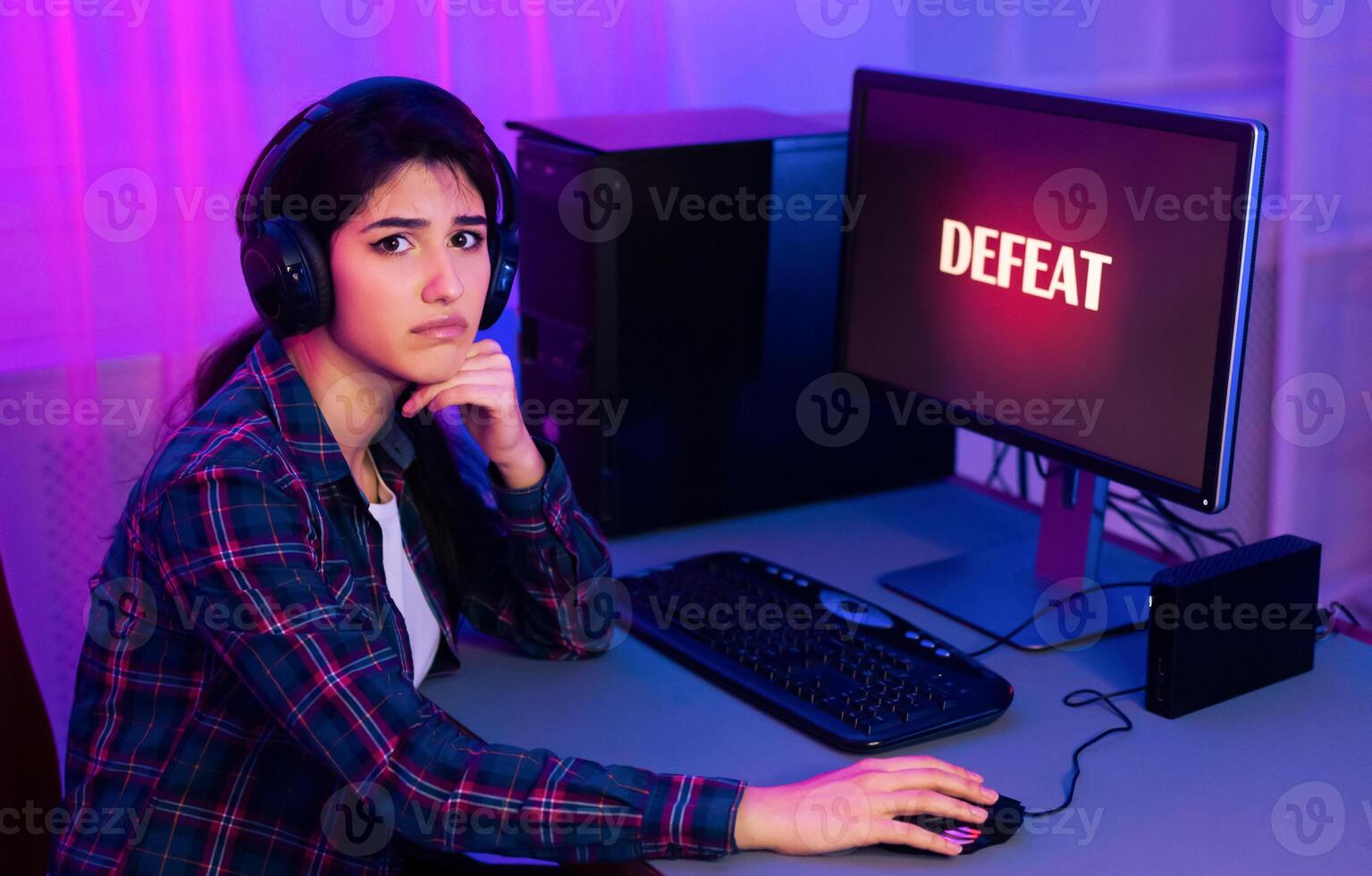 A young woman, wearing headphones, sits at a computer desk with a serious expression on her face. The word DEFEAT is displayed on the computer screen, indicating that she has lost a game photo