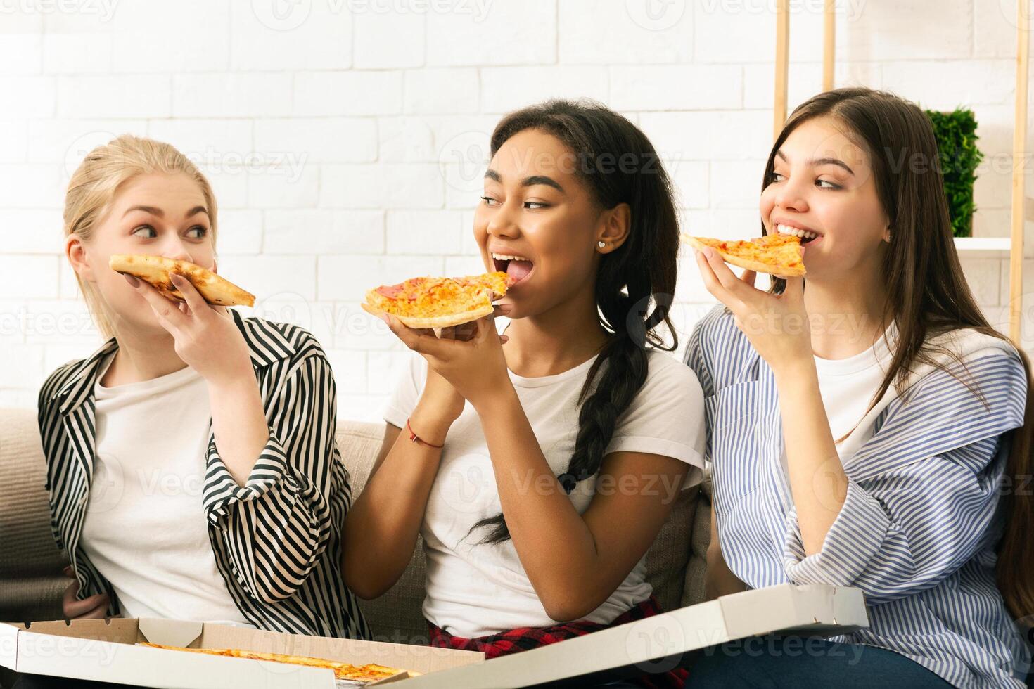 Three young girls are seated on a couch, enjoying slices of pizza. The girls are casually dressed and appear to be having a relaxed and enjoyable time together. photo