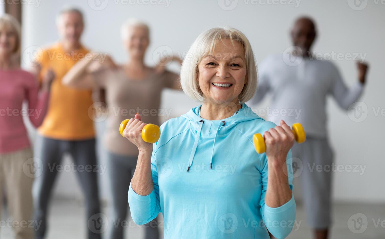 Multiracial group of senior people in sportswear doing strength building fitness exercises with dumbbells, holding fitness tools and smiling at camera, selective focus on positive elderly lady photo