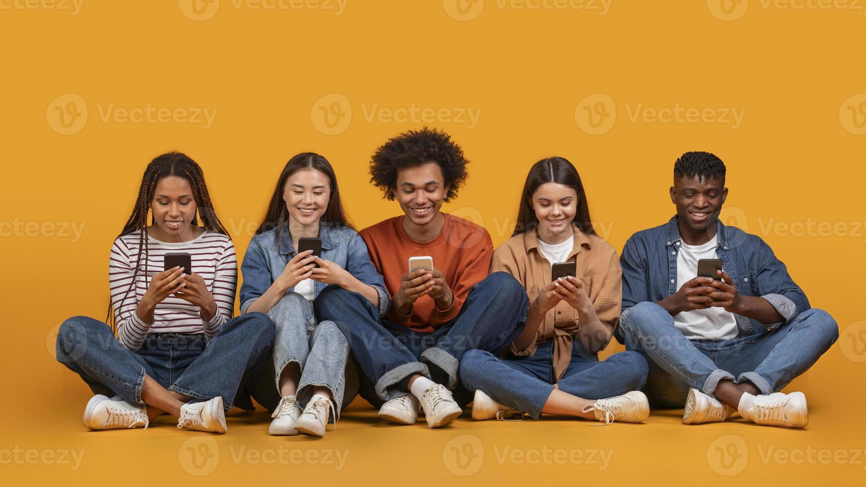 This image features a diverse group of five young adults, all smiling and looking down at their smartphones. They are sitting cross-legged on a yellow background photo