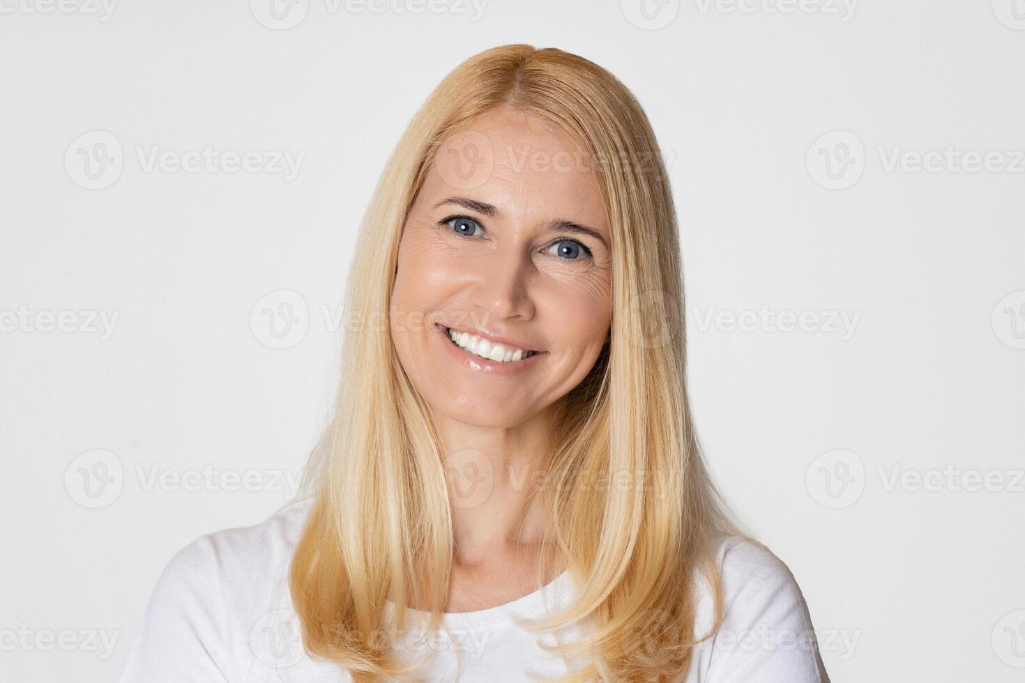 The image is a portrait of a middle aged woman with long blonde hair. She is smiling and looking directly at the camera. She is wearing a white shirt and standing in front of a white background photo