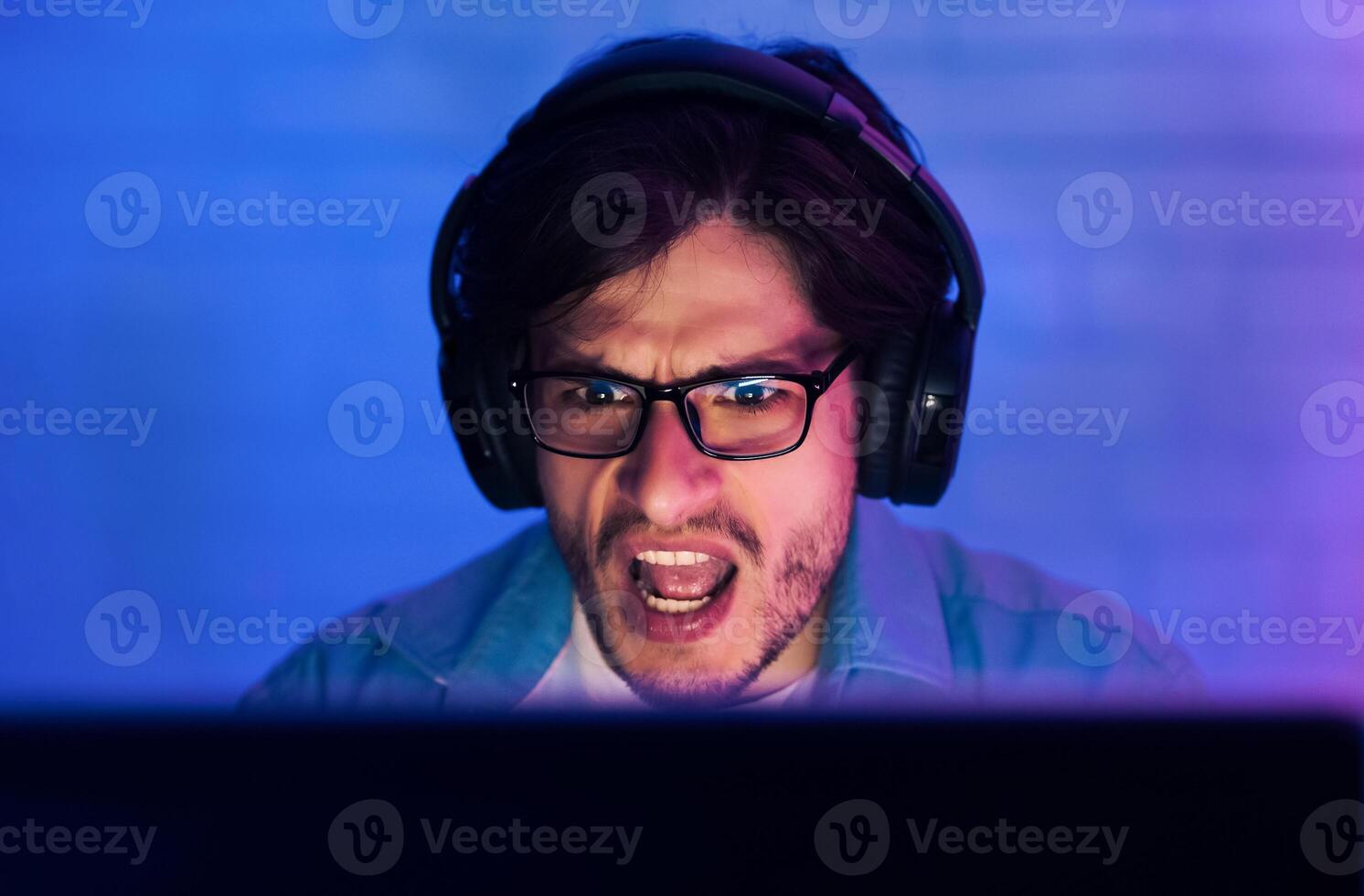 A young man is shown intensely focused on a computer screen, wearing headphones and glasses. His mouth is open in a shout, likely reacting to an exciting moment in a game. photo
