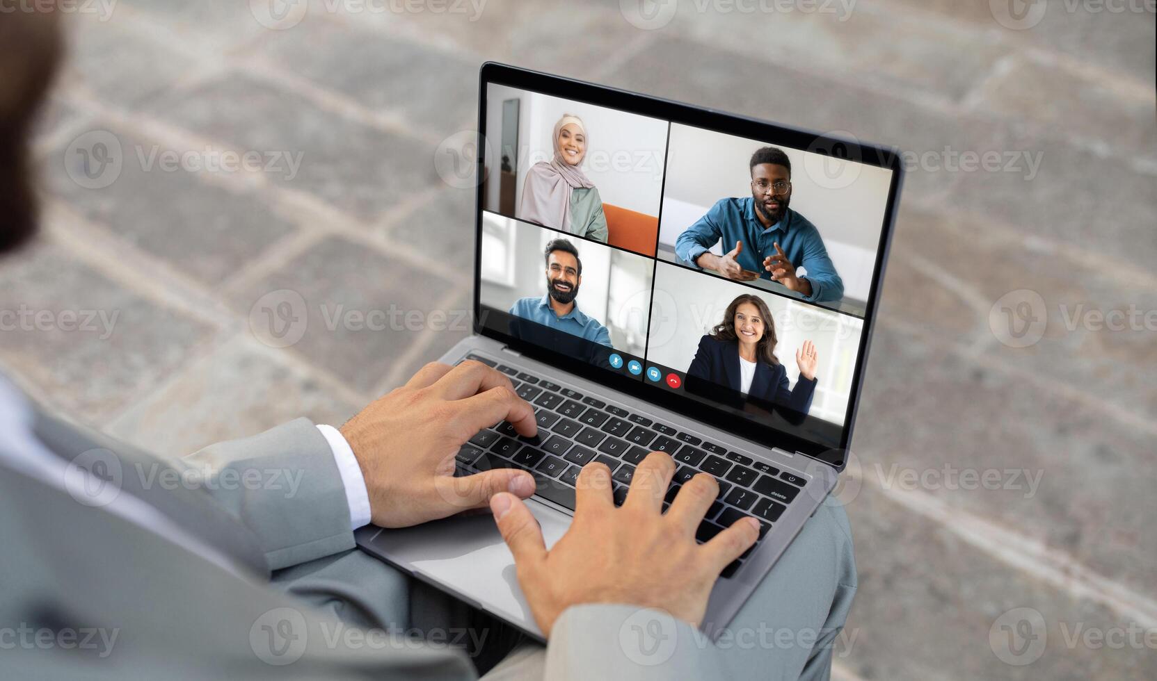 A businessman sitting outside on stone floor is engaged in a team conference call on his laptop. Four diverse colleagues are visible on the screen, actively participating in the virtual meeting photo