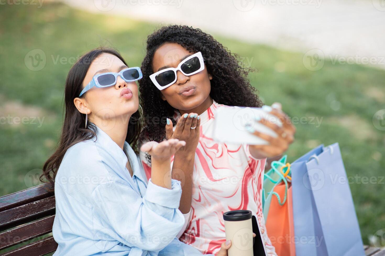 This image shows two multiethnic women taking a selfie in a park. They are holding shopping bags and smiling at the camera. photo