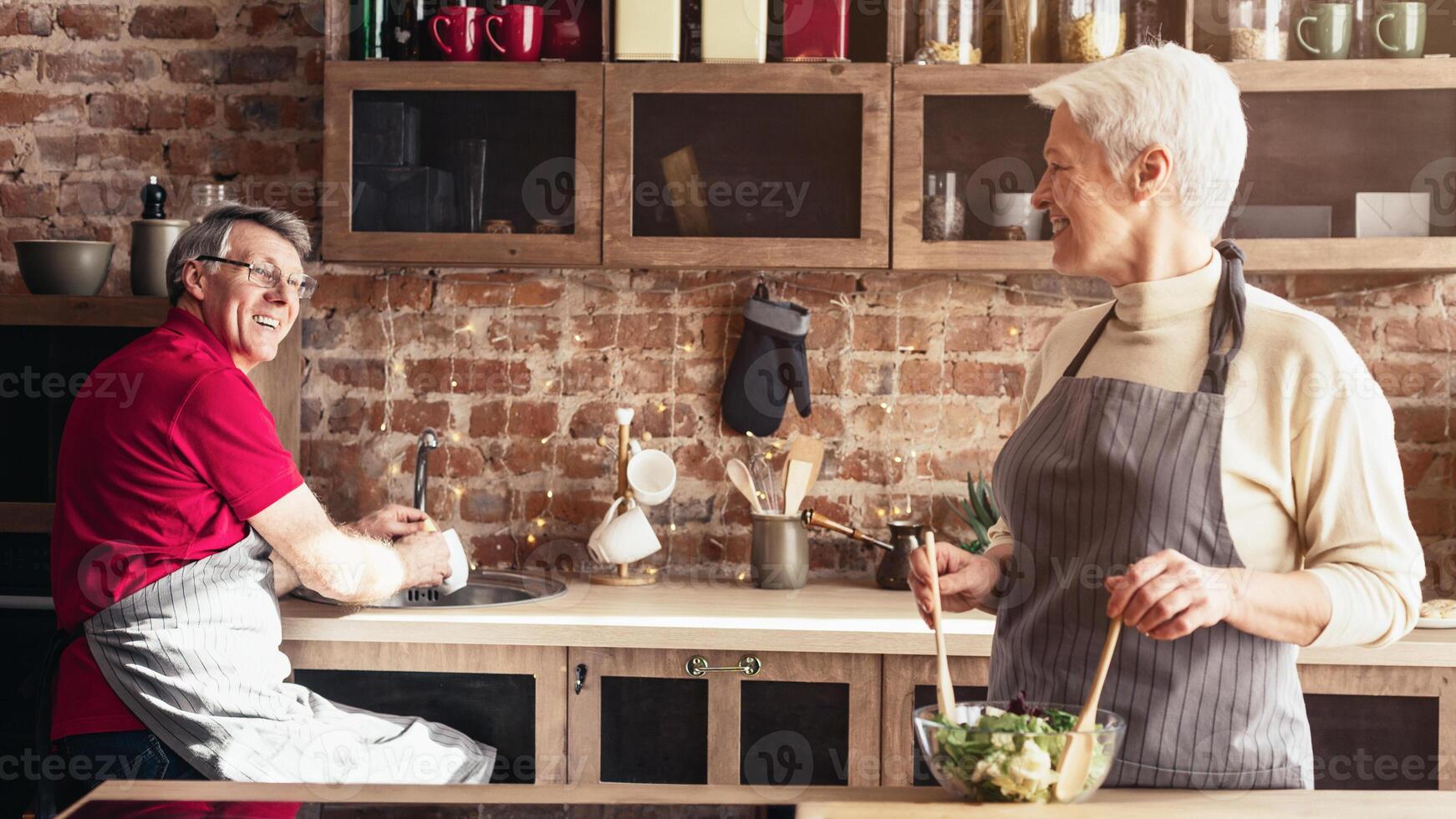 A senior couple is cooking together in a modern kitchen. The man is chopping vegetables, while the woman is standing by the stove. They are both wearing aprons and smiling. photo