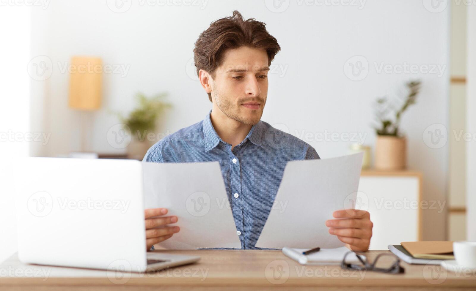 A man in a blue shirt sits at a desk, reading documents. There is a laptop in front of him, along with a notepad and glasses. photo