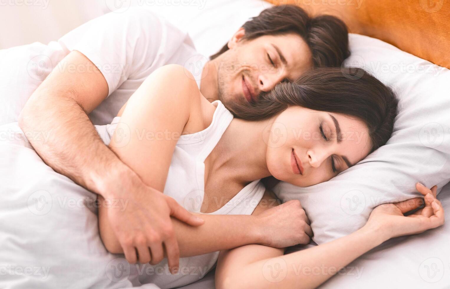 A couple sleeps peacefully in bed together at home. The woman is lying on her side, her head resting on a pillow, while the man is hugging her from behind. They both have their eyes closed photo