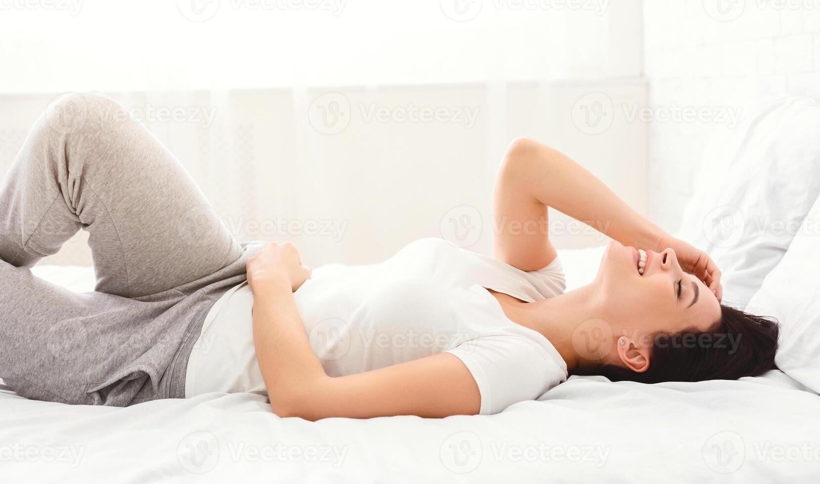A young woman is lying comfortably on a bed with white linens, wearing casual grey sweatpants and a white t-shirt. She appears relaxed and happy, enjoying the sunny morning light photo