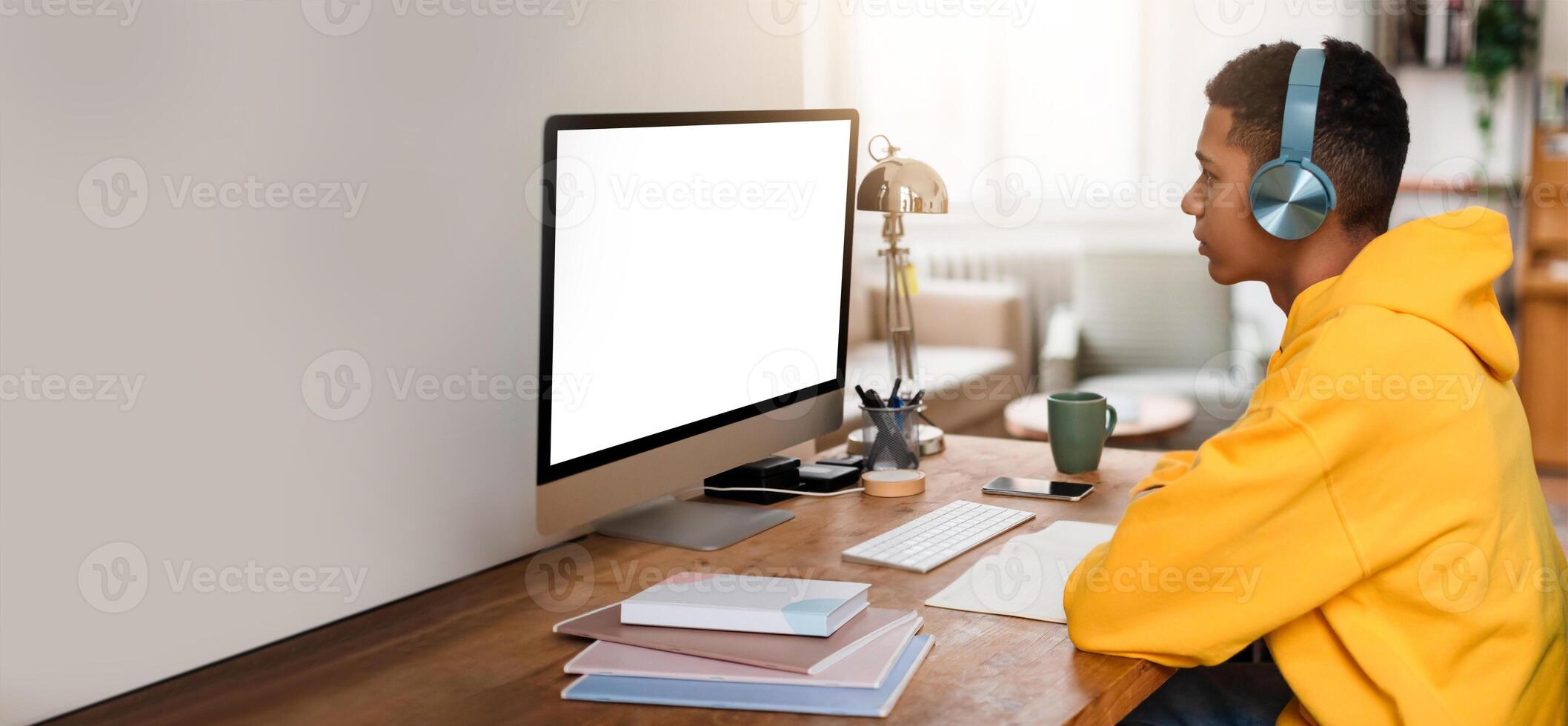 A young black man in a yellow hoodie is intently studying at a wooden desk. He wears headphones while focused on a blank computer screen, surrounded by study materials in a well-lit room. photo