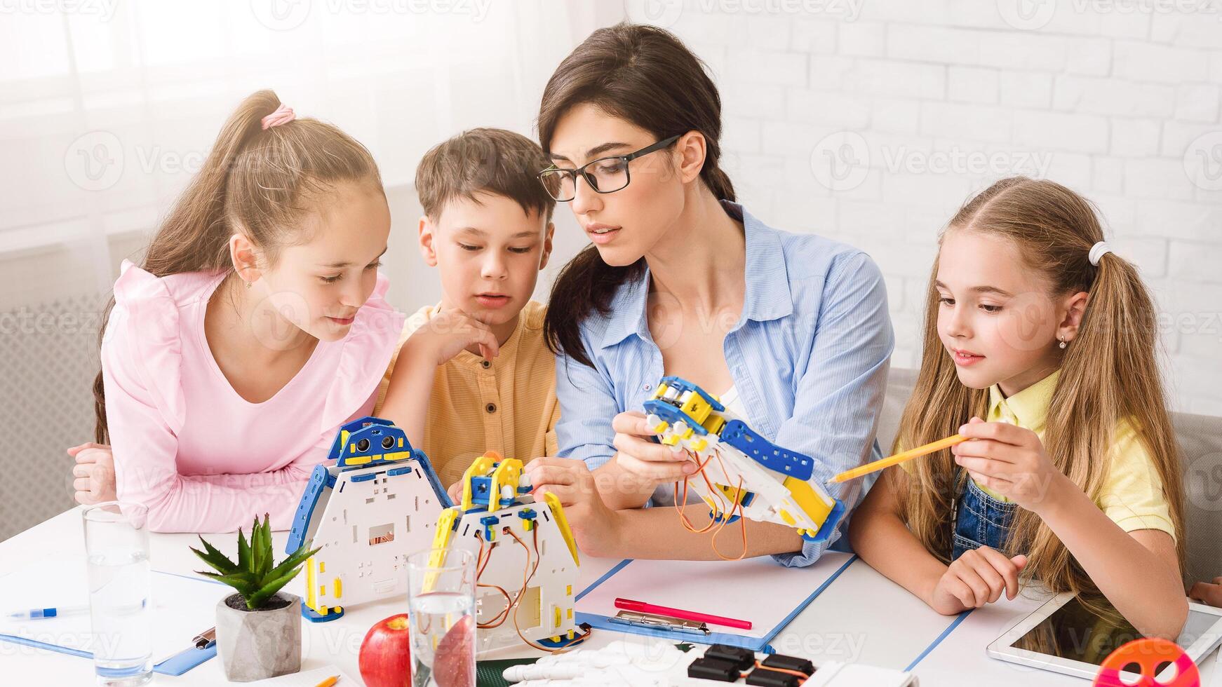 A young female teacher guides three children, two girls and one boy, as they build a robotic arm in a classroom setting. The children are focused and engaged in the activity. photo
