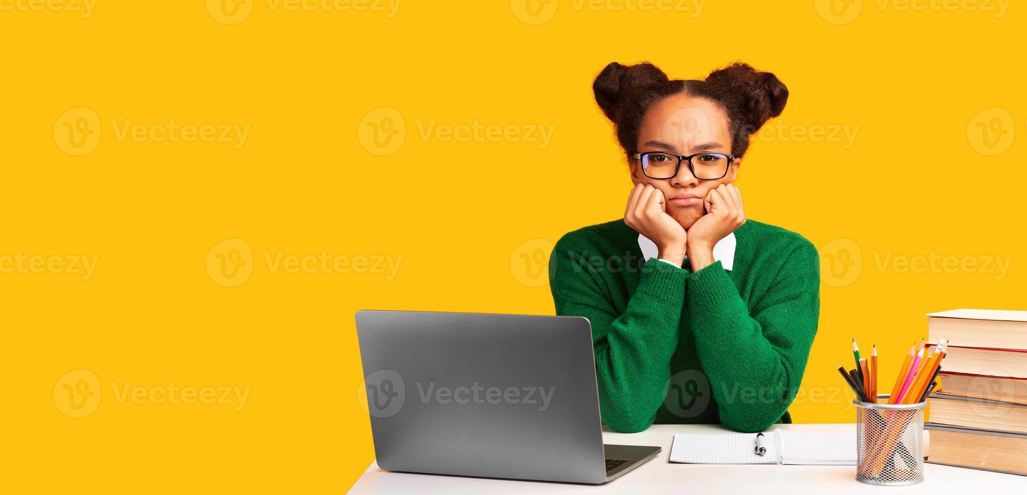 Bored Teenager. Portrait of sad black girl using laptop sitting at table isolated on yellow wall background, copy space photo