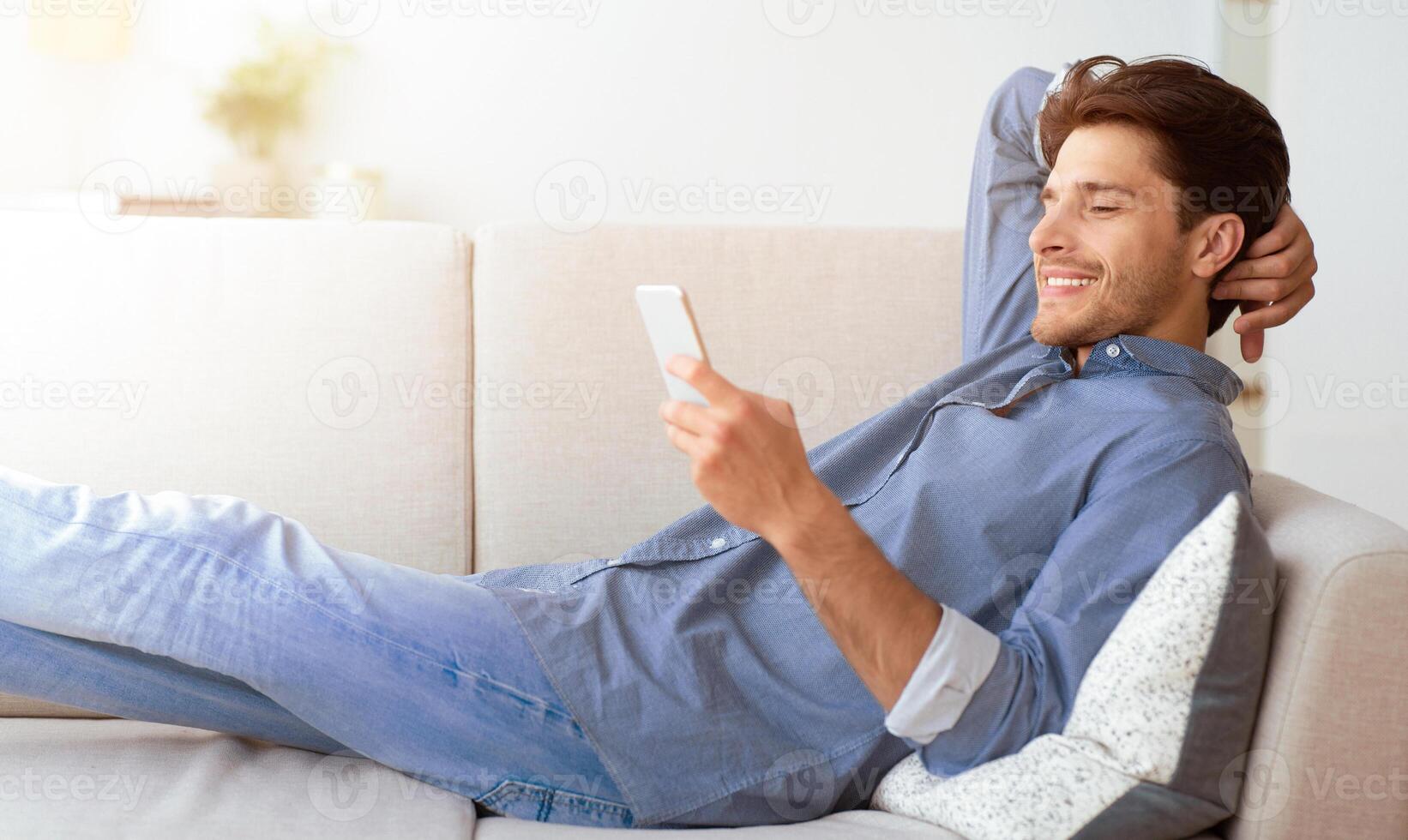 A man is relaxing on a couch, using his smartphone. He is smiling and appears to be enjoying himself. photo
