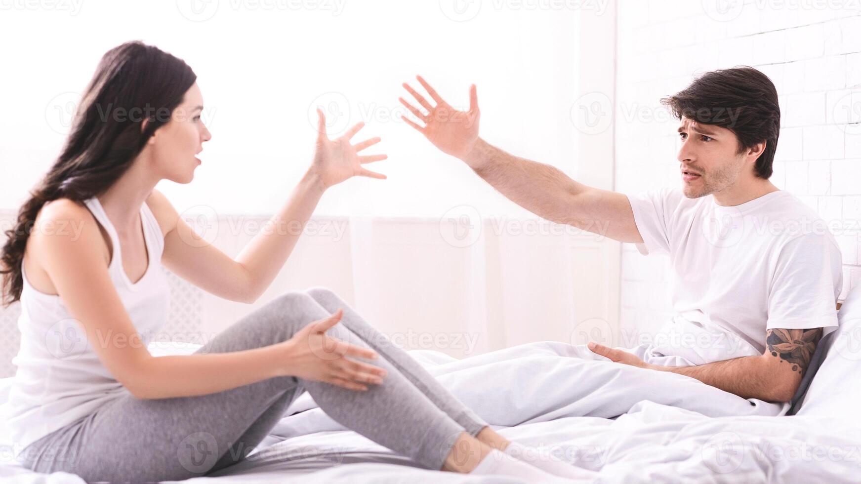 A young couple sits on a bed, engaged in a heated argument. They are both wearing casual clothing and appear to be upset. The image suggests a tense and possibly contentious relationship. photo