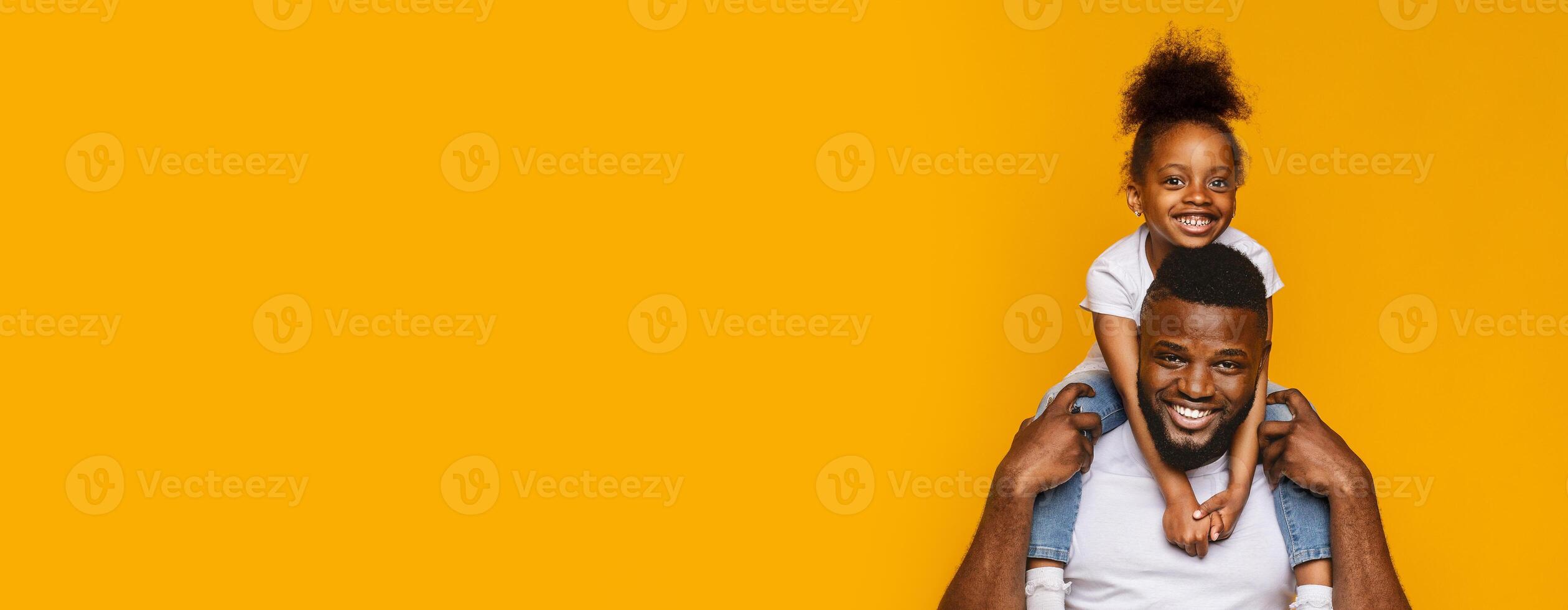 A smiling black father carries his cheerful daughter on his shoulders, both sharing a moment of joy and laughter. The vibrant yellow background enhances the warmth of this loving interaction photo