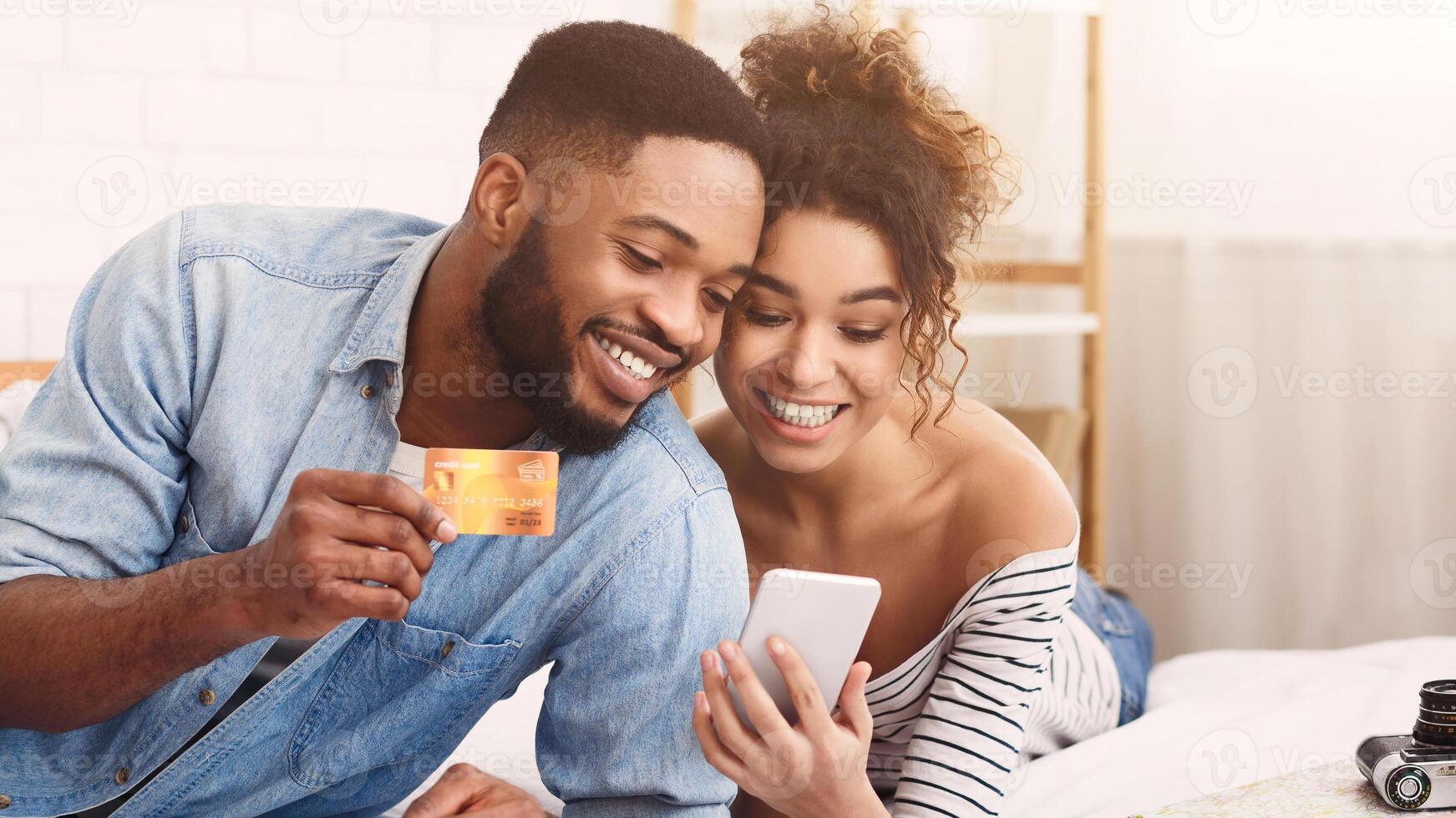 African American cheerful young couple sits closely together as they make an online purchase using a mobile phone and credit card. photo