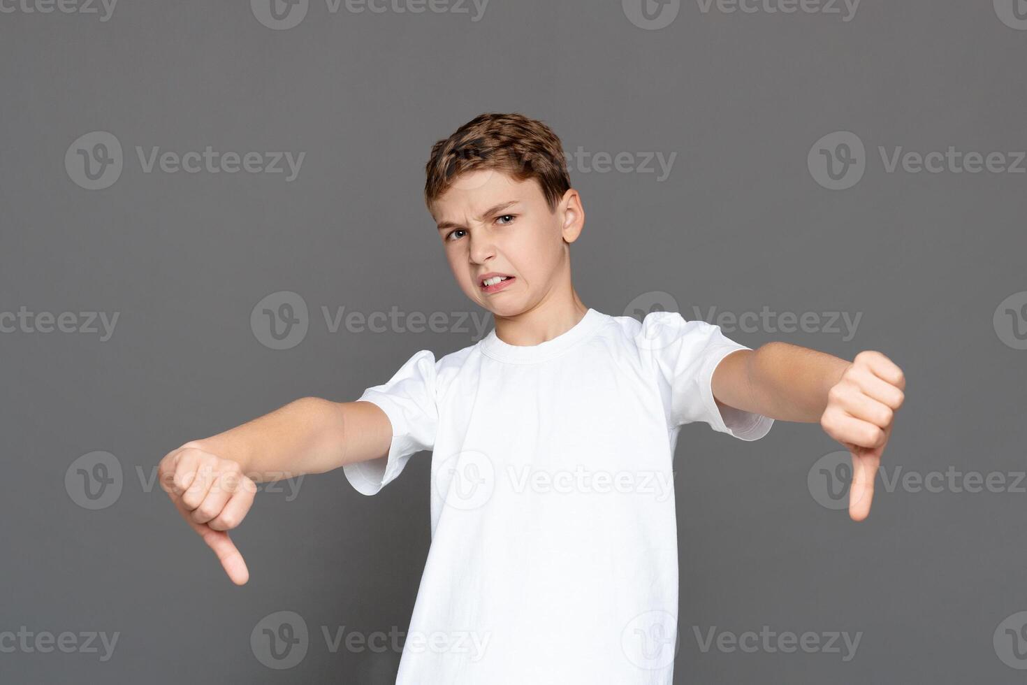 A young boy stands against a grey background, expressing dissatisfaction with both hands showing thumbs down photo