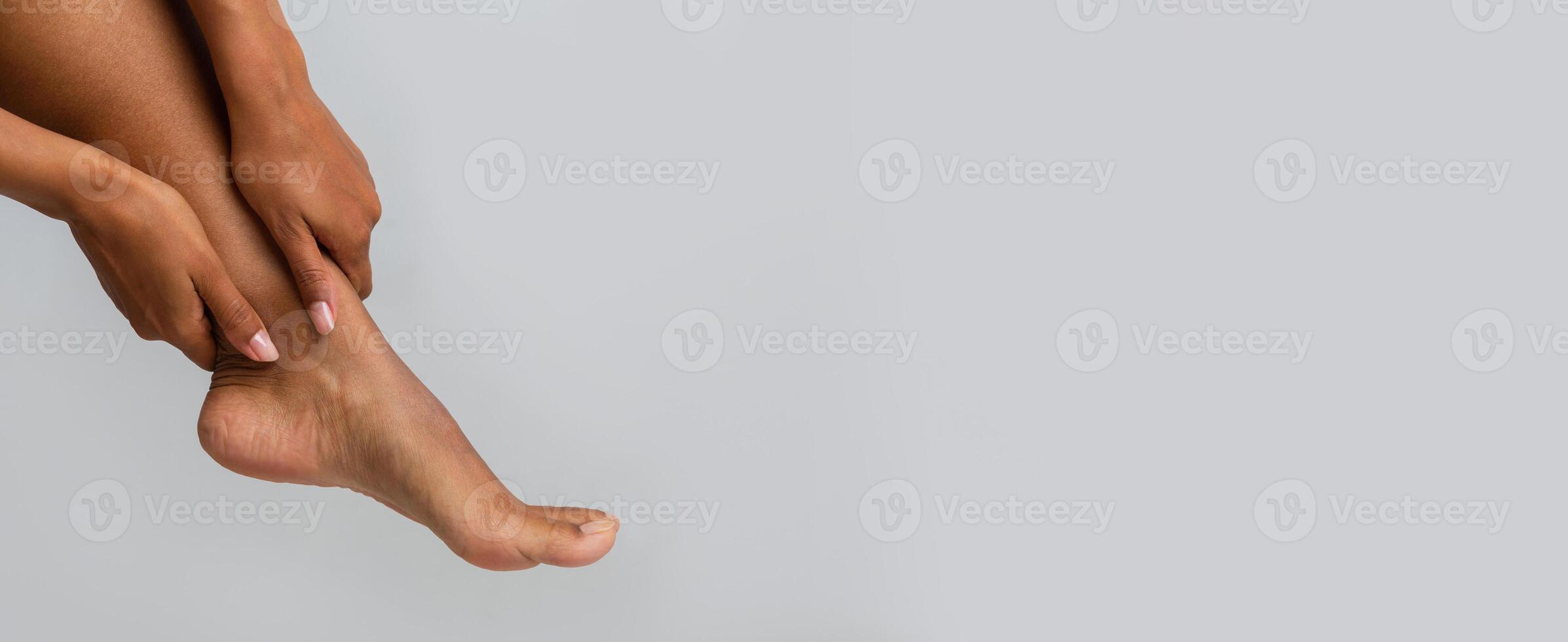 Cropped of afro woman rubbing her injured ankle over grey background, panorama with copy space photo