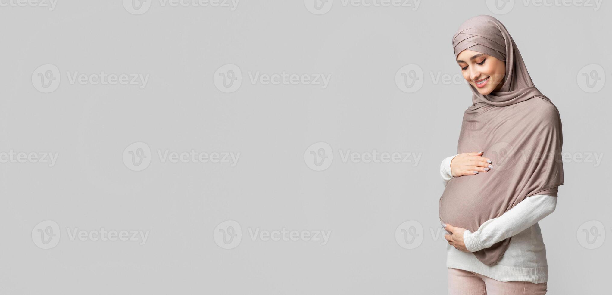 Enjoying pregnancy. Pregnant muslim woman in headscarf tenderly touching her belly, embracing tummy over gray background with empty space, panorama photo