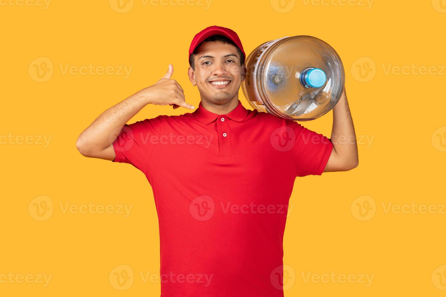 Delivery man wearing a red shirt is holding a huge bottle of water in his hand and showing thumb up on yellow studio background photo