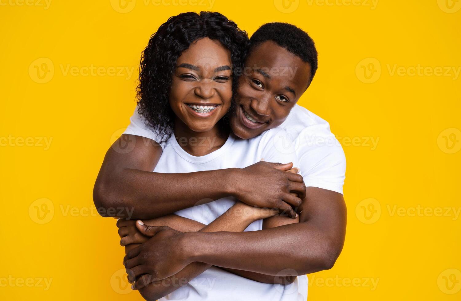 Loving black couple cuddling over yellow studio background. Cheerful young african american man and woman hugging and smiling at camera. Love, relationships, marriage concept photo