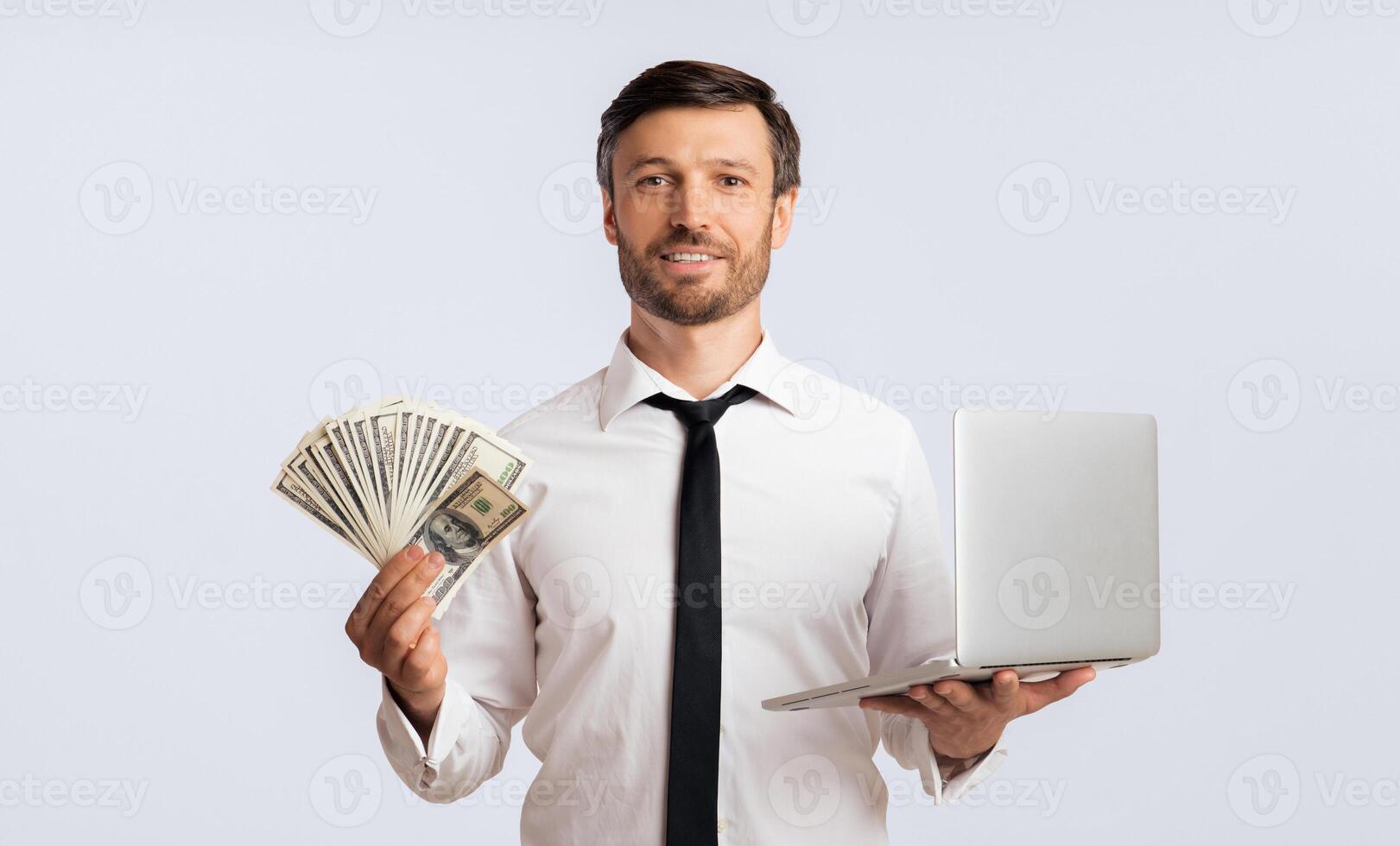 A smiling man in a white shirt and tie holds a fan of cash in one hand and a laptop in the other, against a white background. photo