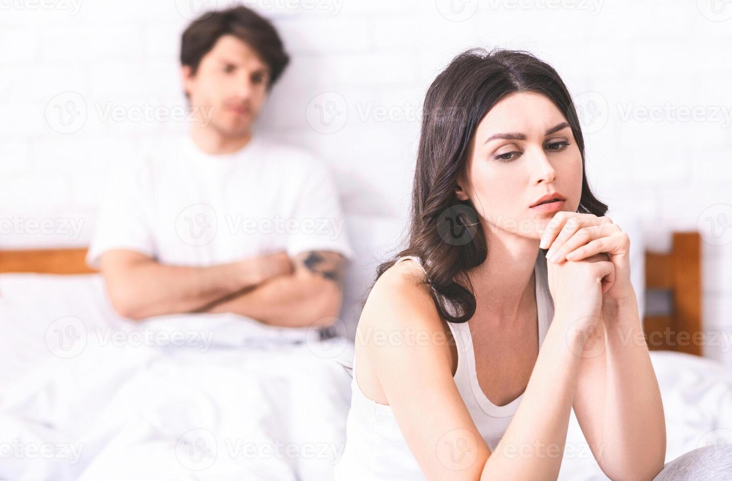 The image shows a woman sitting on a bed, looking sad and lost in thought. She is wearing a white tank top and has her hands clasped together in front of her. photo