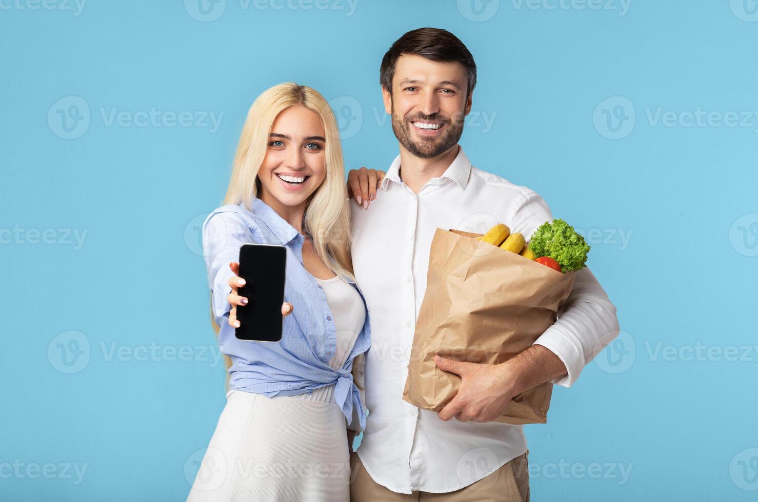A young woman and man stand together, the woman holding a smartphone out toward the camera and the man holding a paper bag full of groceries. They are both smiling and appear to be happy. photo