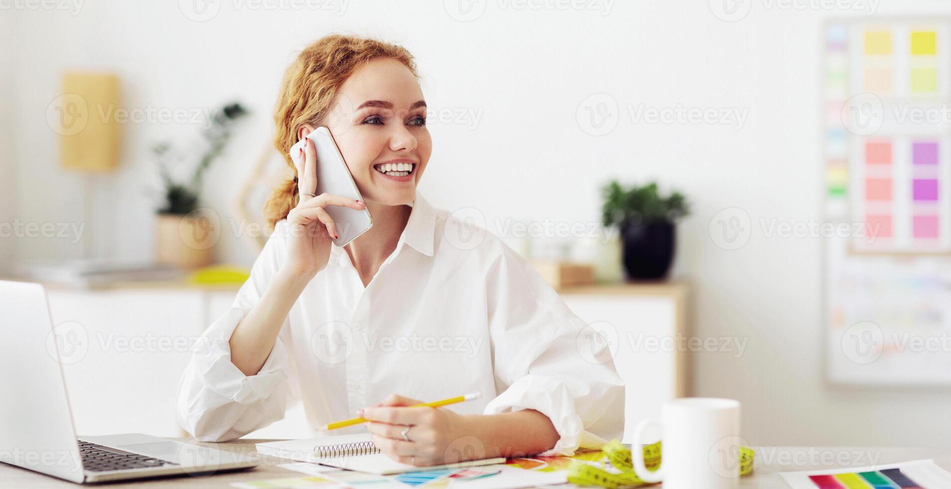 A woman smiles and talks on her phone while sitting at her desk. She is working on a project, with a laptop and other materials in front of her. photo