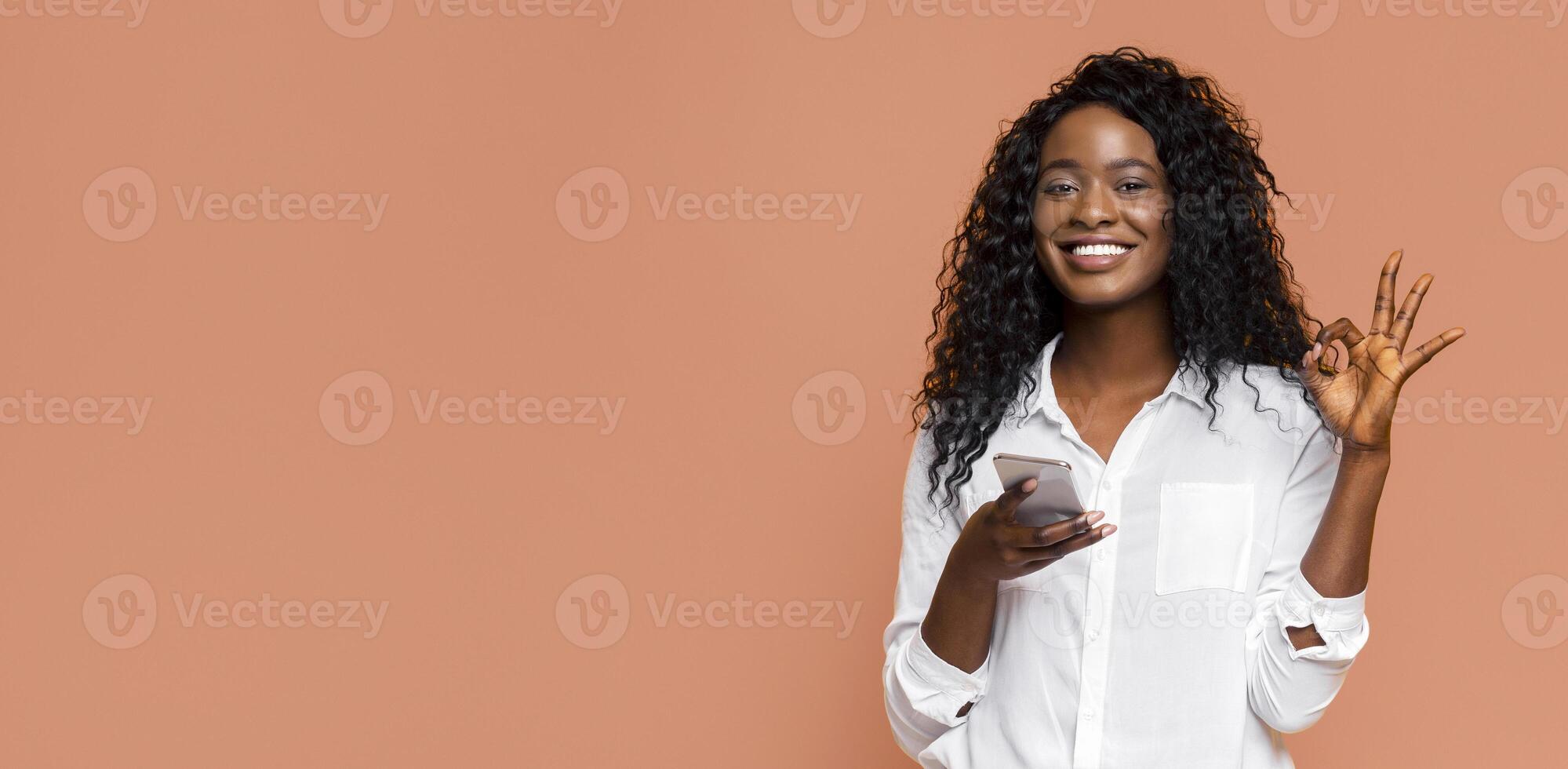 A young black woman with long, curly black hair is wearing a white button-down shirt. She is smiling and making the ok sign with her right hand. She is holding a white smartphone in her left hand. photo