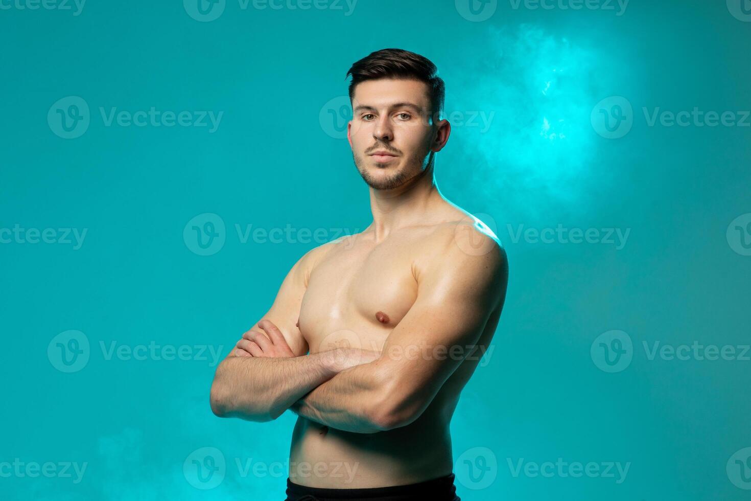 A confident young man stands shirtless with arms crossed, looking directly at the camera. The background is a vibrant blue, accentuated by fog effects, creating a dramatic and striking atmosphere. photo