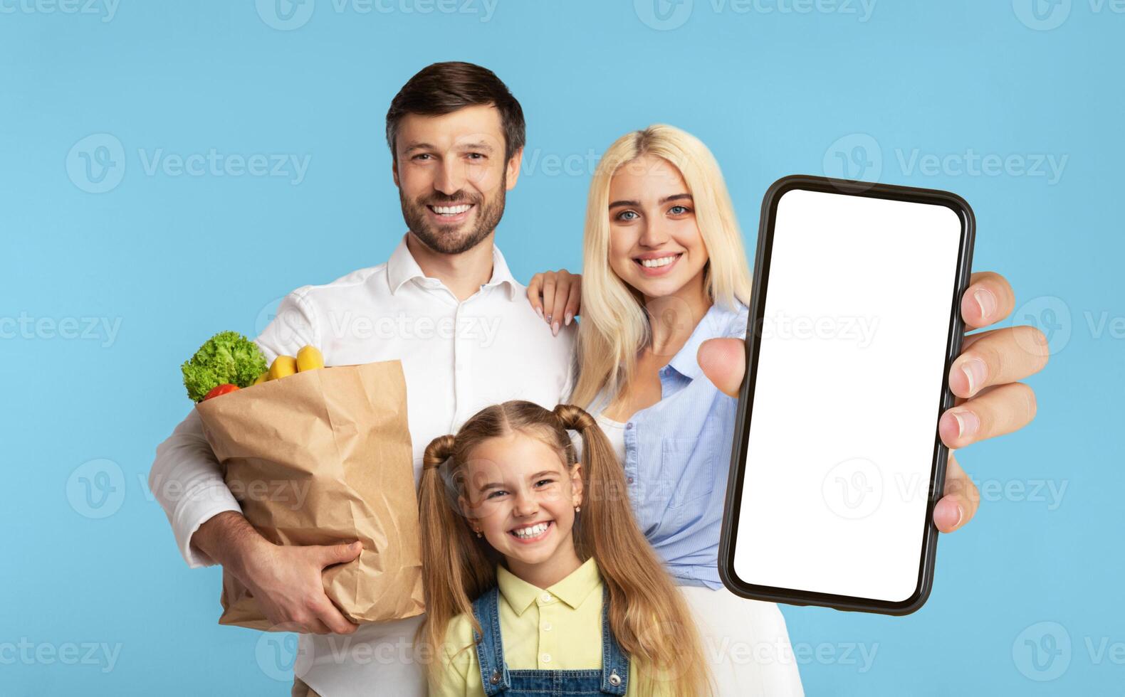 A happy family of three stands in front of a blue background. The mother holds a smartphone with a blank screen towards the camera. The father holds a paper bag full of groceries. photo