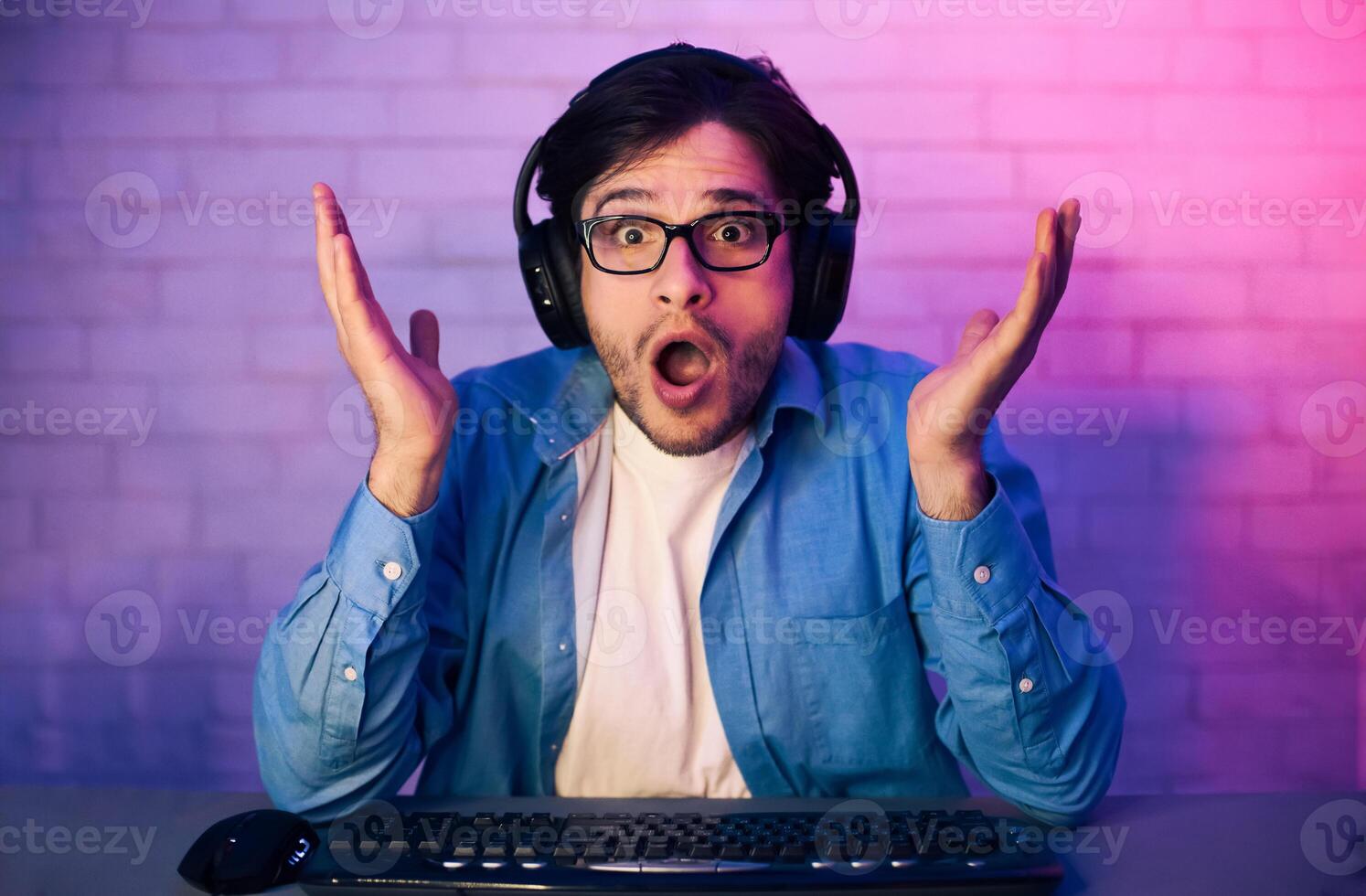 This image features a young man, wearing headphones, sitting in front of a computer and looking shocked and surprised. He is wearing a blue shirt and appears to be playing a game. photo