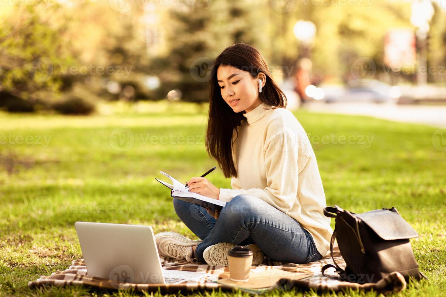 Asian woman sitting on green grass with a laptop and notebook open in front of her, appearing to be working or studying outdoors. photo