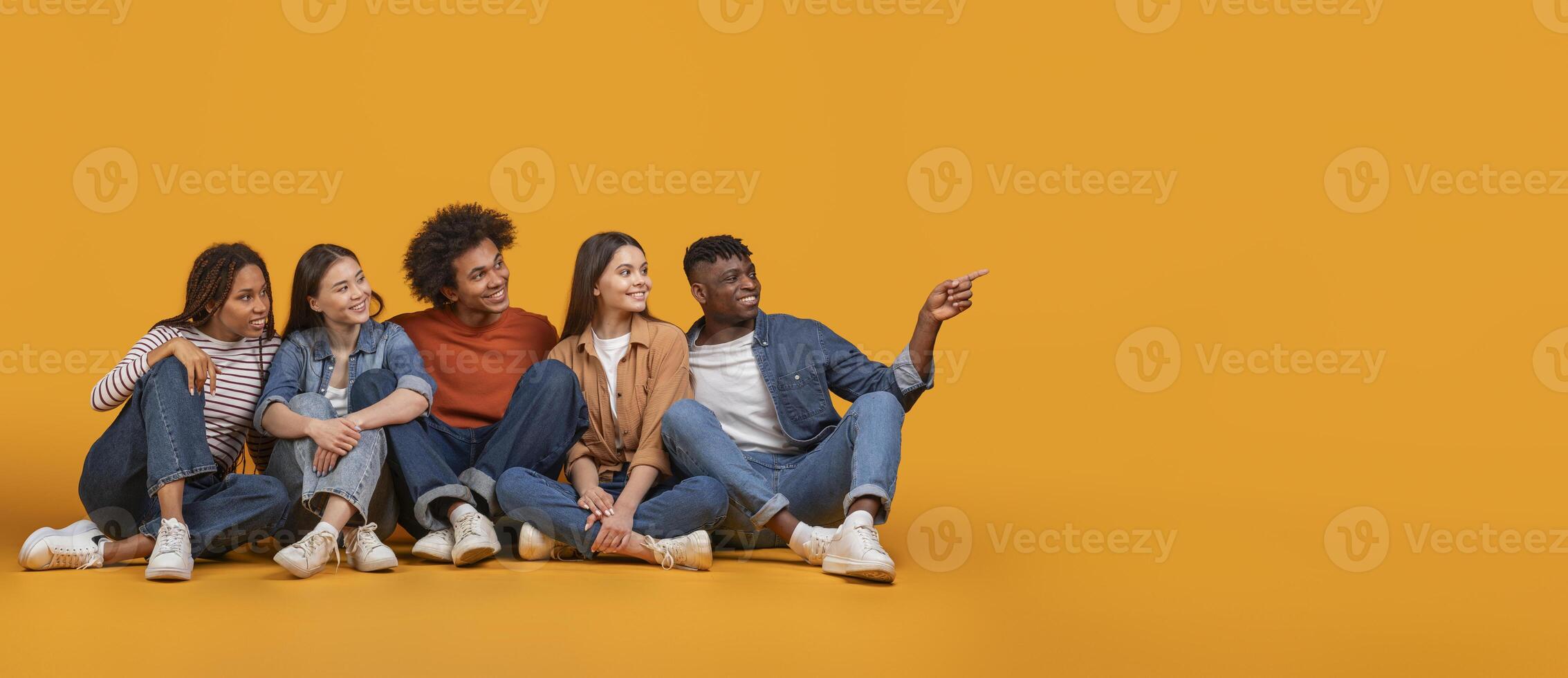 Five young adults, a diverse group of friends, are sitting on a yellow background. They are all smiling and looking to their right, with one person pointing towards the empty space photo