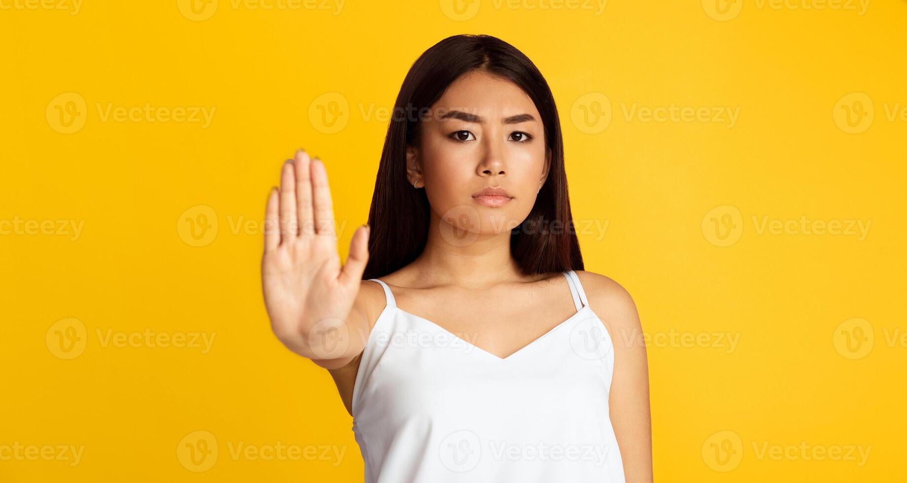 A young Asian woman stands against a bright yellow background. She is facing the camera and holding out her left hand with her palm facing forward in a stop gesture photo