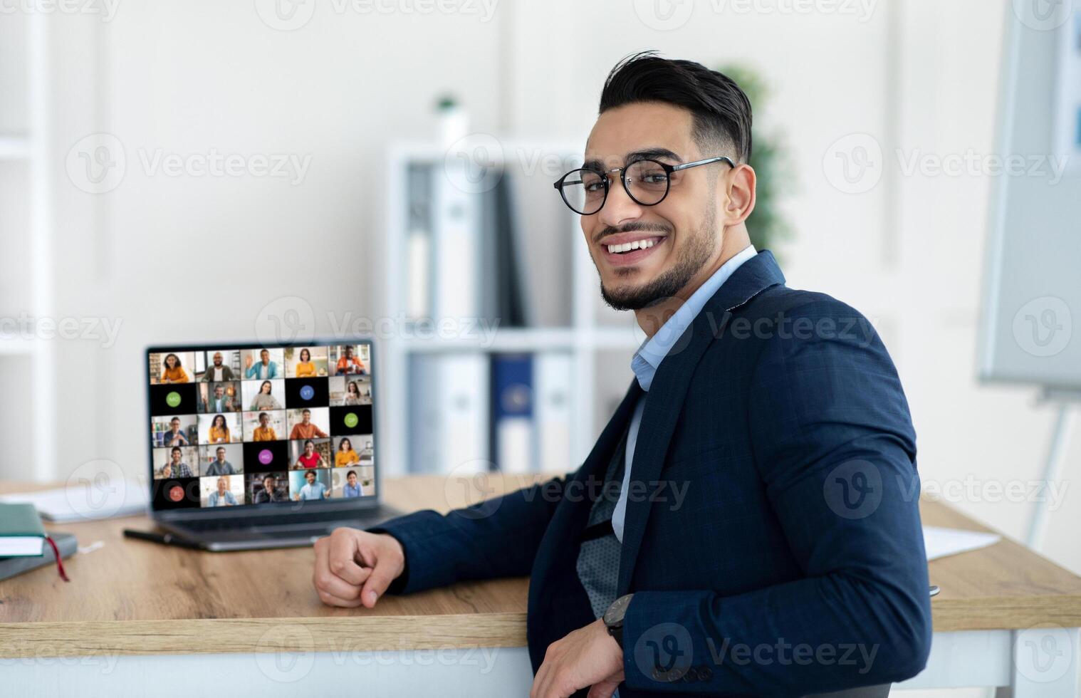 A young middle eastern businessman wearing glasses sits at his desk in an office, smiling as he looks away from his laptop. The laptop screen displays a conference with many participants photo