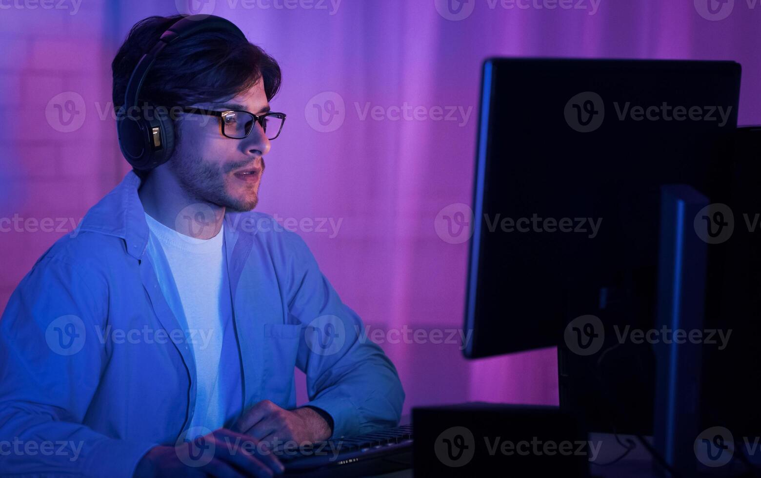 A young man with headphones and glasses is sitting at a computer in a room lit by purple and blue lights. He is looking at the monitor with a focused expression. photo